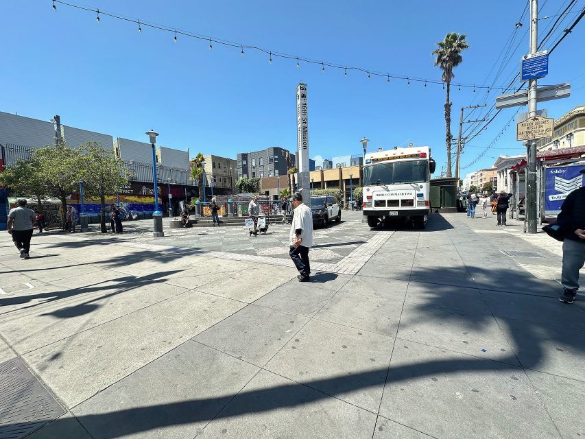 A city street scene with pedestrians walking, a parked white truck, string lights overhead, and buildings in the background under a clear blue sky.