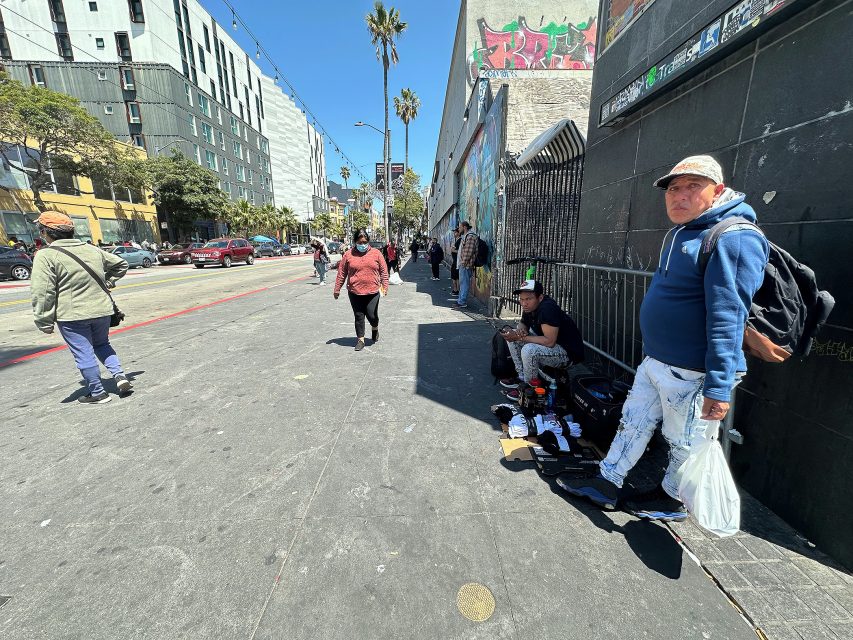 People walk along a sunlit urban sidewalk lined with buildings, graffiti, and palm trees; some individuals stand or sit by the wall with bags and belongings.