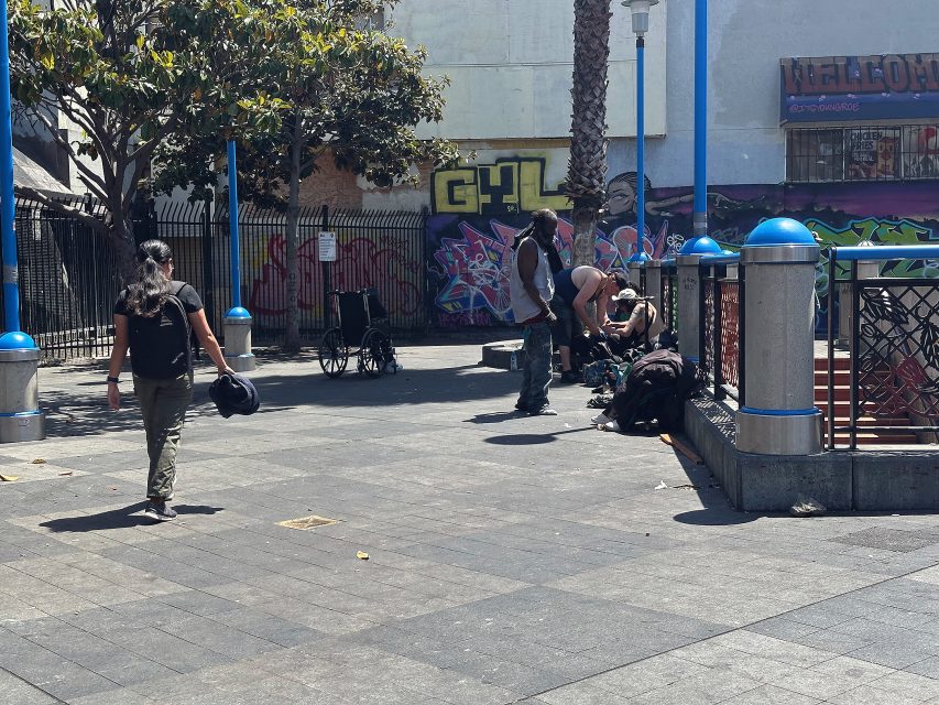 A person walks across a plaza while a group of people gather near a fenced area with graffiti and a wheelchair in the background.