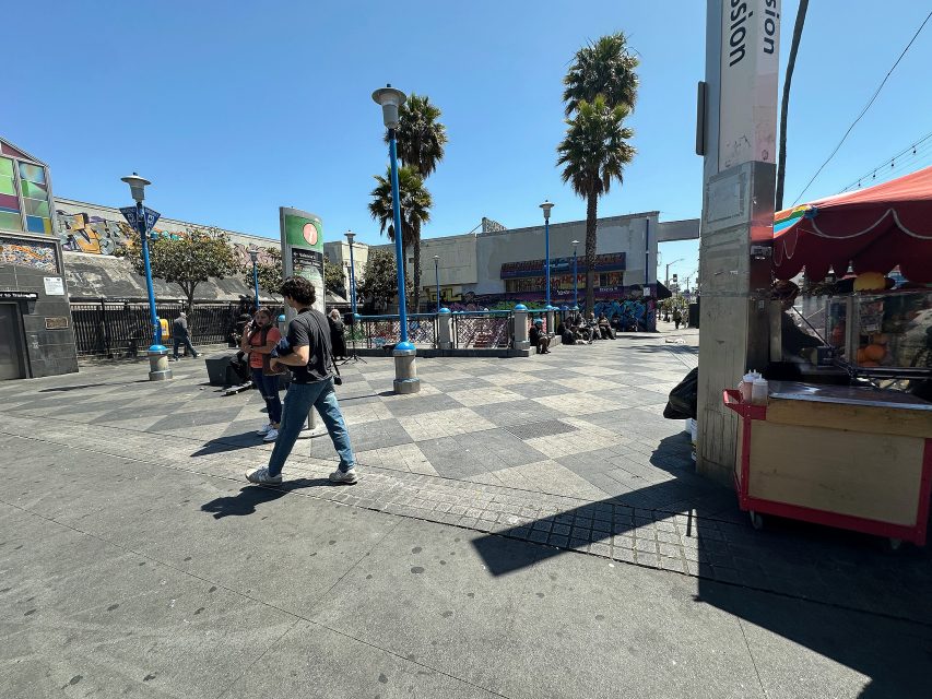 People walk through a sunny urban plaza with palm trees, a food cart, murals, and market stalls in the background.