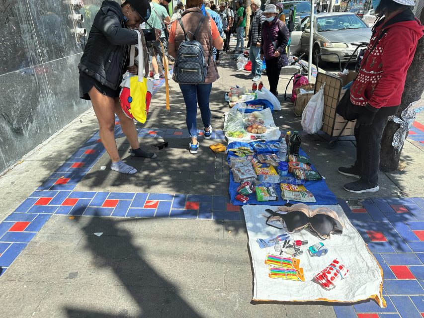 People browse various items, including food, toiletries, and clothes, laid out on blankets and tarps for sale on a city sidewalk.