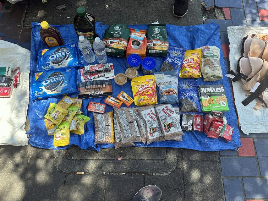 Assorted packaged food items, drinks, and toiletries displayed on a blue tarp on the ground, with bras and other items on a separate cloth to the right.