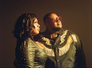 Two people stand side by side, both wearing dark outfits with gold accents, looking slightly upward into the distance against a plain background.