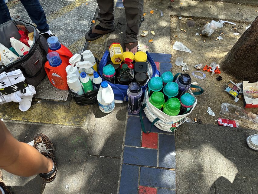 Bottles of cleaning products and plastic tumblers are displayed on the sidewalk, surrounded by litter and people's feet.