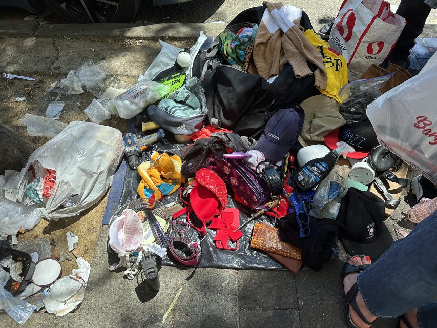 A pile of assorted personal belongings, clothing, tools, plastic bags, and miscellaneous items scattered on a sidewalk.