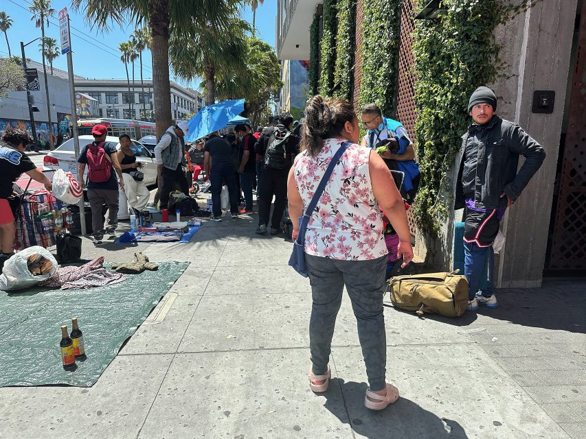 A group of people gather on a sunny city sidewalk, some with belongings or blankets, and others standing or conversing near a building.