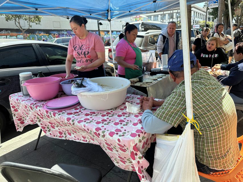 Two women prepare food behind a table covered with large bowls under a canopy at a busy outdoor market while people sit and eat nearby.