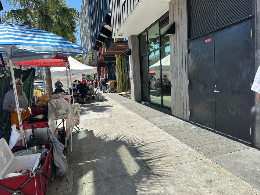Outdoor market with vendor tents on a sunny day; people are sitting and shopping along a sidewalk beside modern buildings.