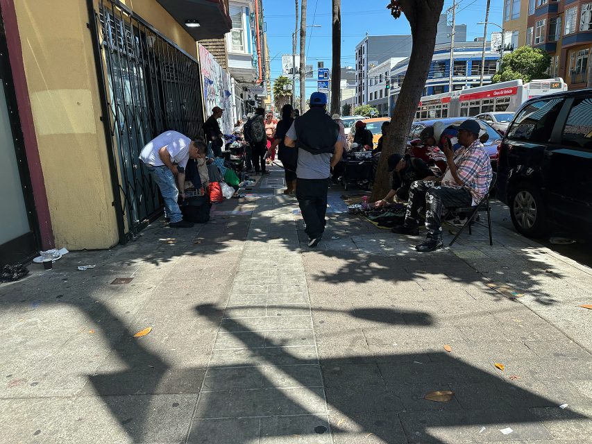 People sit, stand, and gather along a city sidewalk on a sunny day, with buildings, parked cars, and a bus visible in the background.