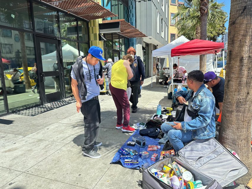 People browse items for sale laid out on blankets and in suitcases on a city sidewalk, next to market stalls and palm trees.