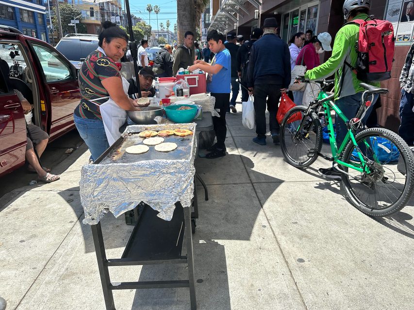 A woman cooks food on a griddle covered with foil on a busy sidewalk as people walk by, and a man with a green jacket stands with a bicycle.