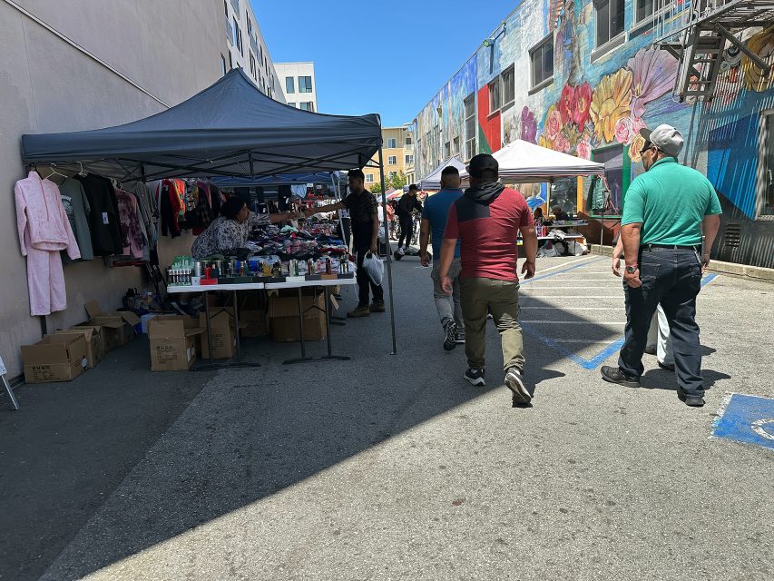 People walk through an outdoor market with vendor tents selling clothes and other goods on a sunny day, with colorful murals on the building walls.