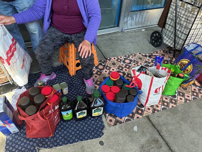 A person sits on a stool outside with bags and containers filled with groceries, including jars, bottles of oil, and packaged goods, arranged on patterned mats.