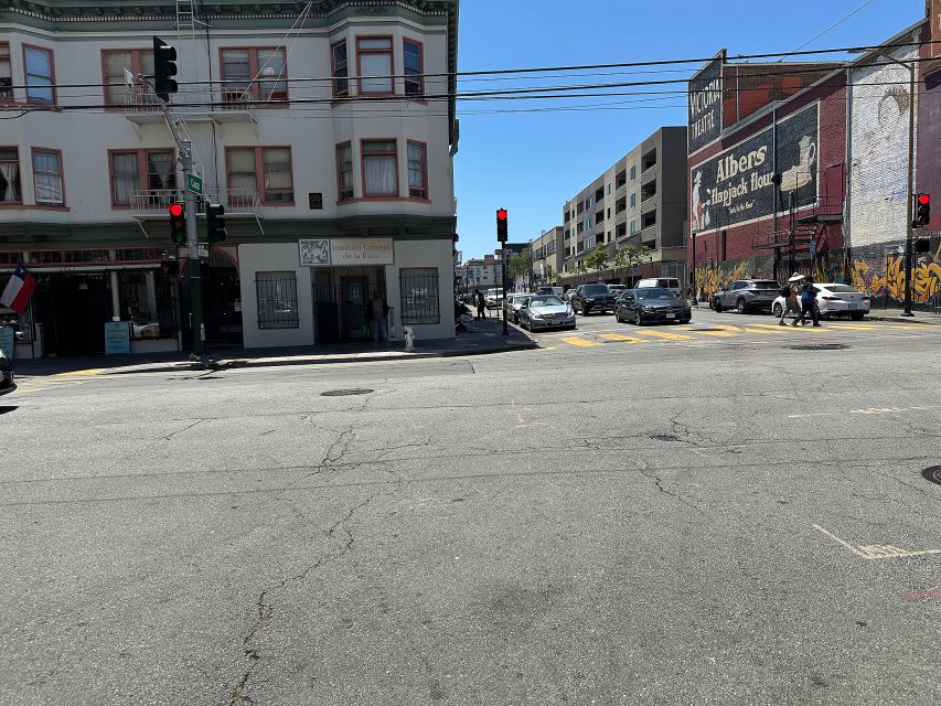 Intersection with traffic lights, cars waiting, pedestrians crossing, and buildings with murals and signs on a sunny day.