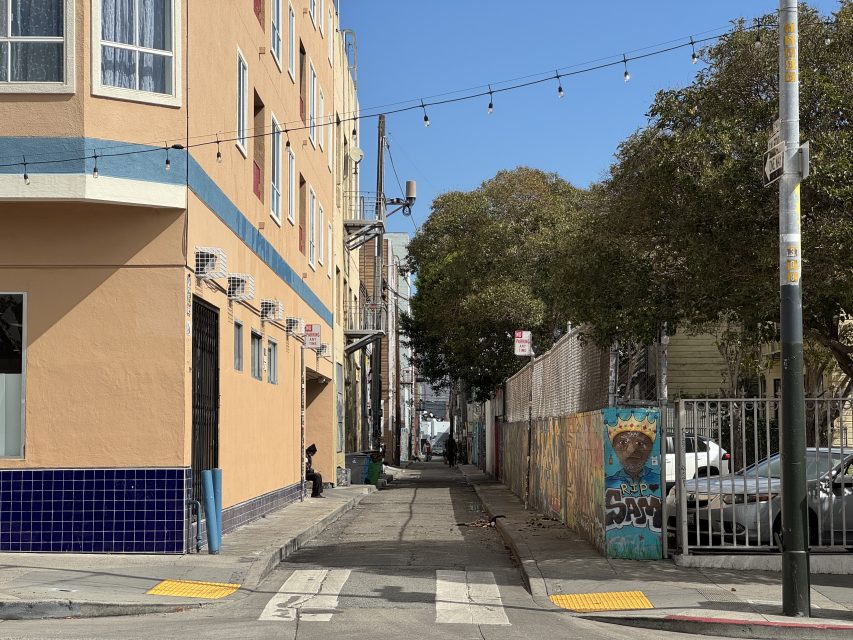 A narrow urban alley lined with a mural-covered fence, trees, a peach building with blue tiles, and overhead string lights under a clear sky.