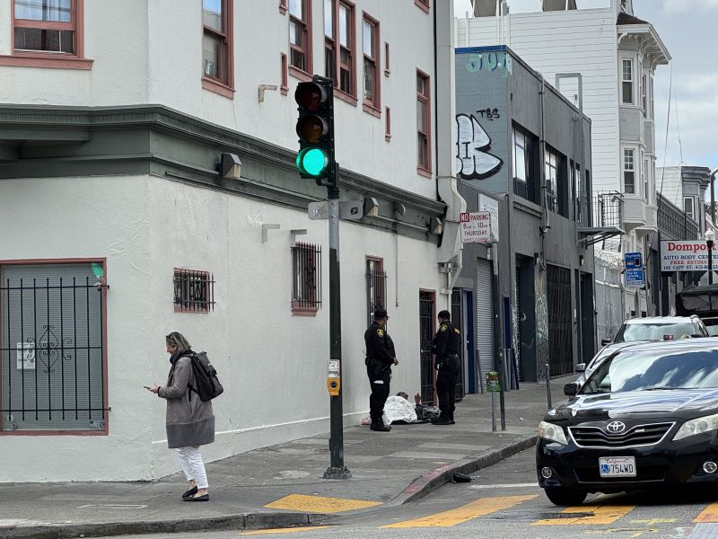 A person walks across a crosswalk while the traffic light is green; two people stand near bags on the sidewalk beside a city building.