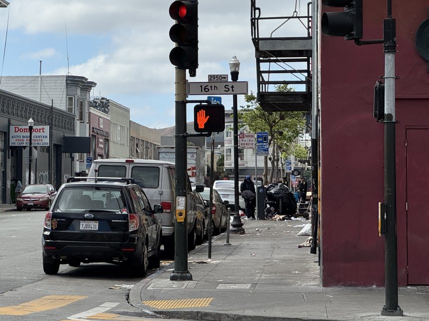 Street view at 16th St intersection with parked cars, a red stop hand pedestrian signal, and people near scattered trash on the sidewalk.