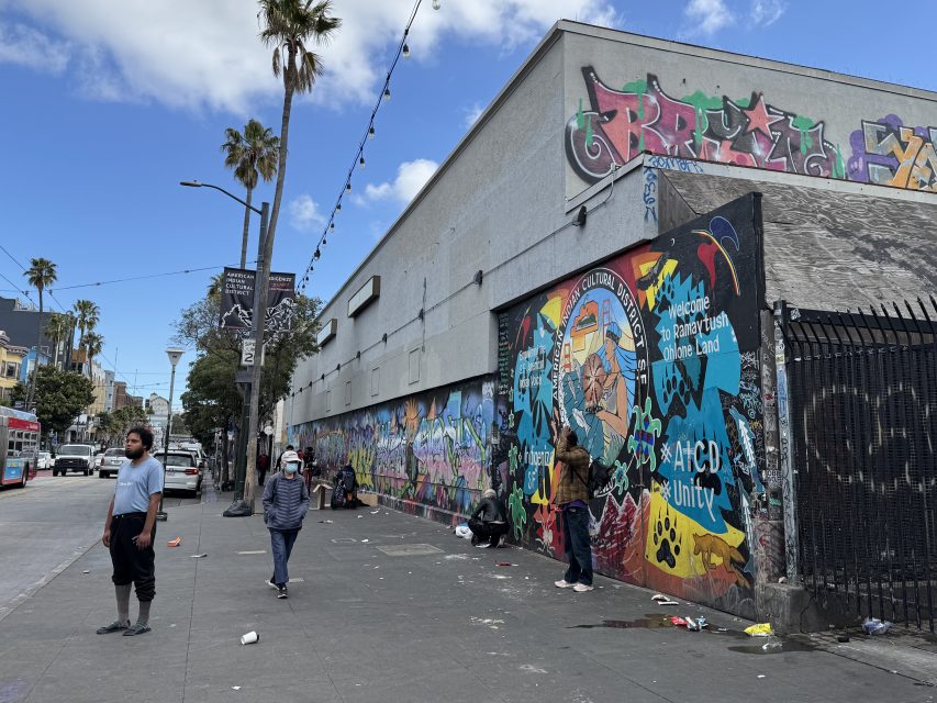 People walk along a city sidewalk beside a building covered in colorful murals and graffiti under a partly cloudy sky.
