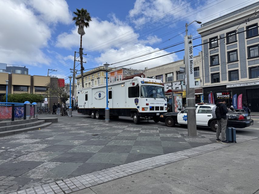 A large white police vehicle is parked on a city street near a BART station entrance, with people walking and a police officer standing nearby.