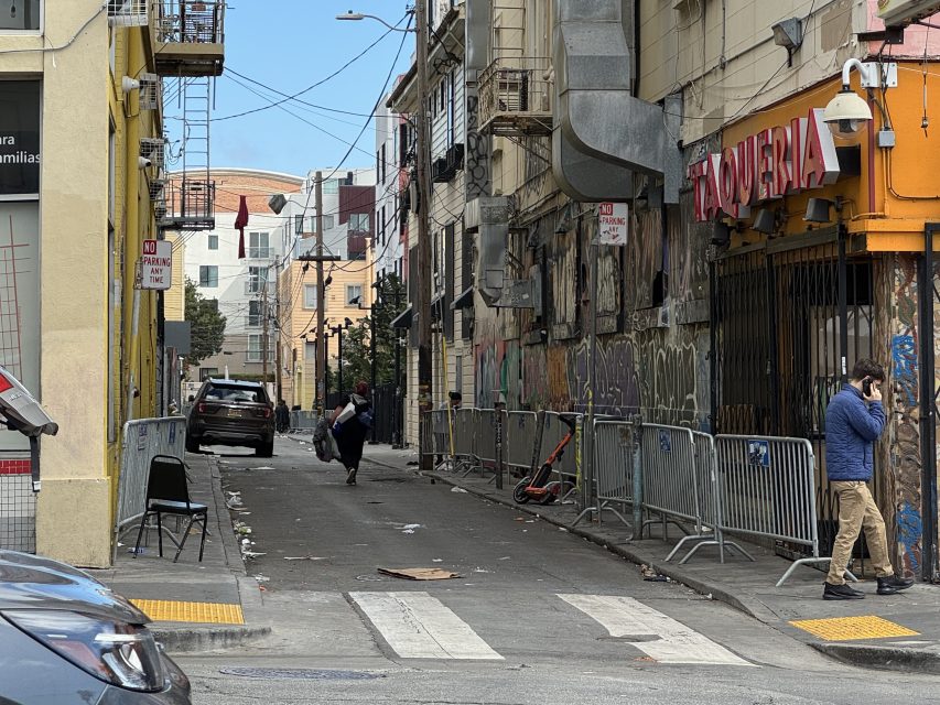 A narrow urban alley with graffiti, metal barricades, and scattered debris. A person sweeps the ground while another stands nearby on a phone. A car is parked along the alley.