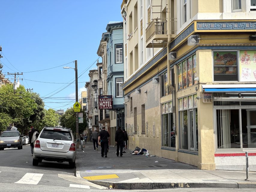 People walk along a city sidewalk beside a hotel and a corner store, with parked cars and a street sign visible under a partly cloudy sky.
