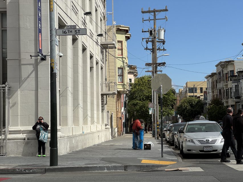 A city street corner at 16th Street with parked cars, pedestrians, power lines, and buildings under a clear blue sky.