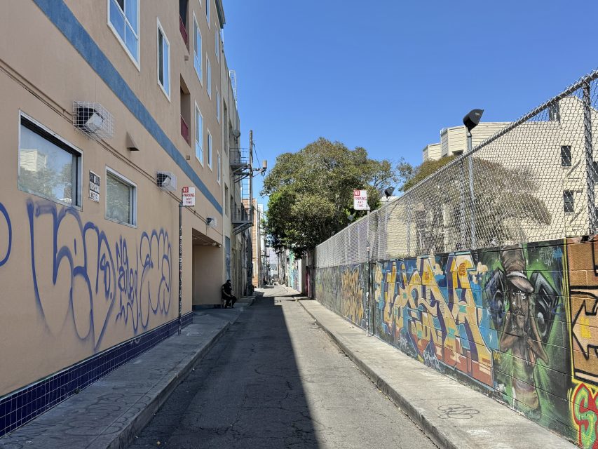 A narrow urban alleyway with graffiti on the building and fence, under clear blue skies, with a person sitting in the distance.