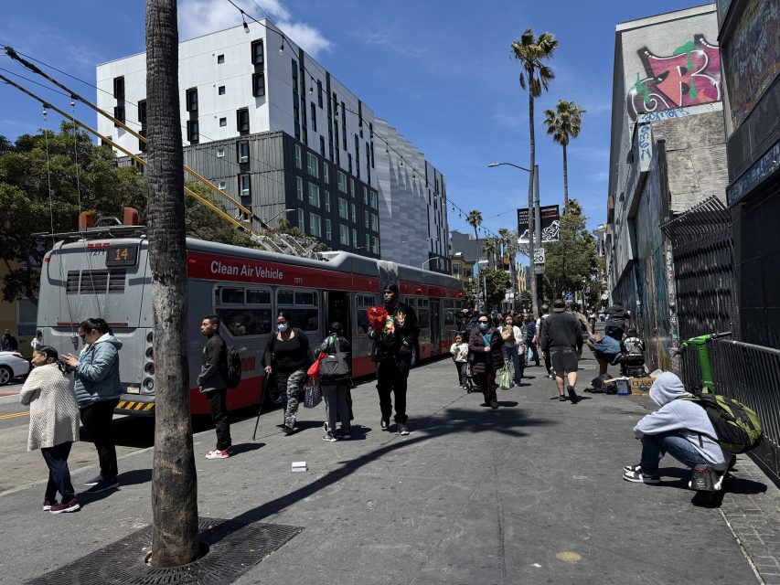 A city street scene shows people waiting near a red bus; some stand, others sit, with tall apartment buildings and palm trees in the background under a partly cloudy sky.