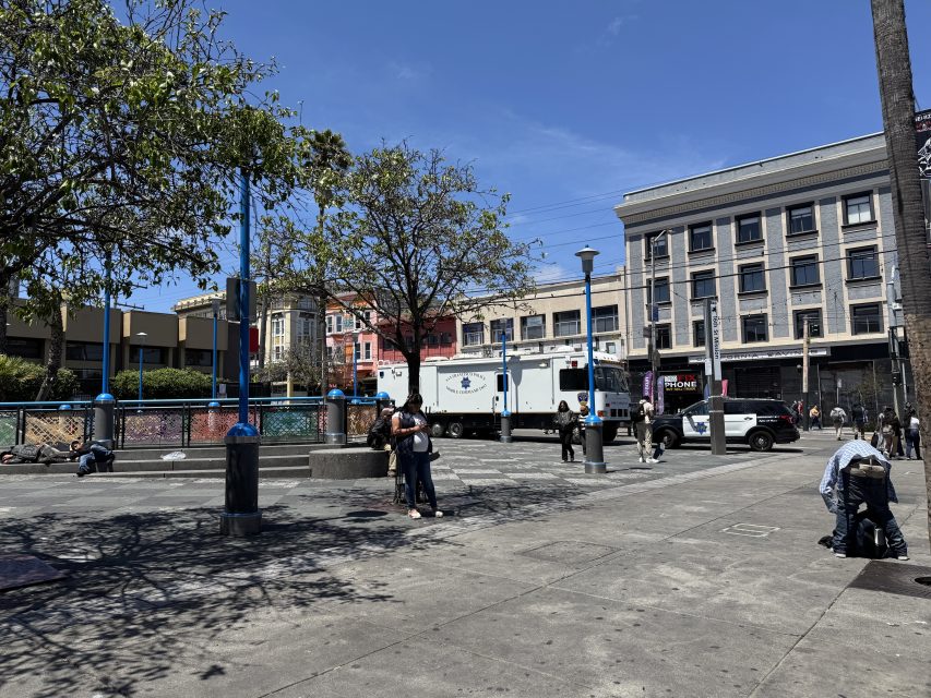 A city square with people walking and sitting; a white police truck and a police car are parked in front of buildings on a sunny day.