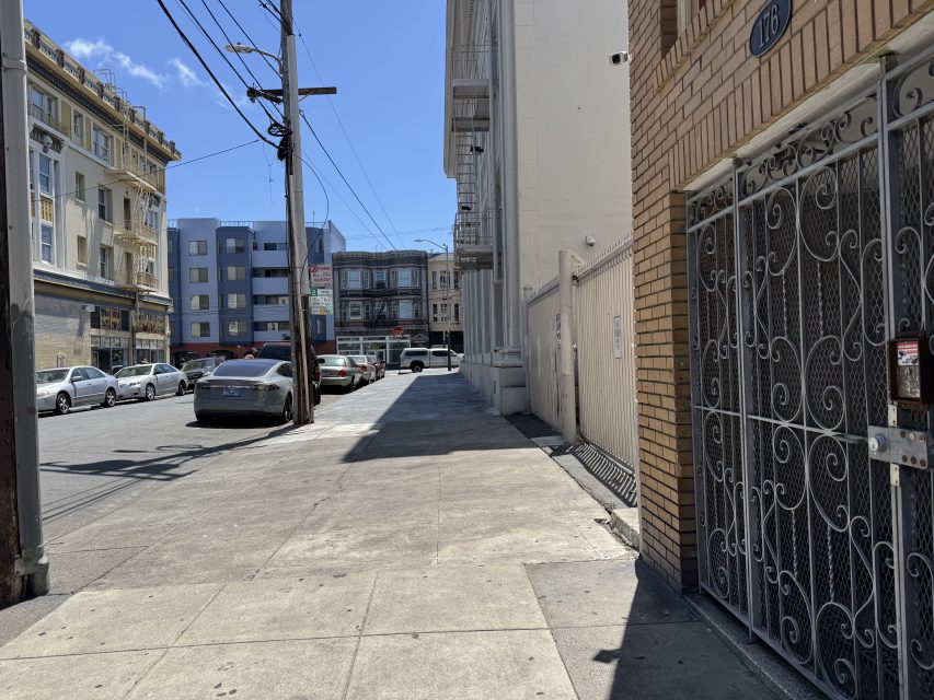 A city sidewalk with parked cars along the street, multi-story buildings on both sides, and a gated entrance in the foreground on a sunny day.