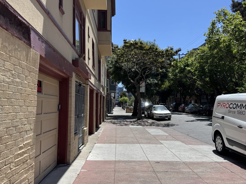 A quiet city sidewalk lined with trees, parked cars, and buildings on a sunny day. A white van is parked on the right side of the street.