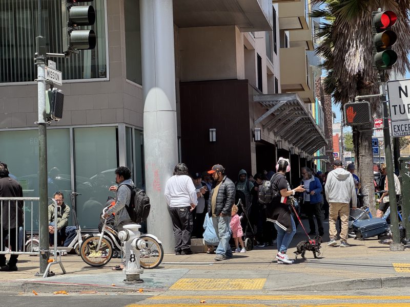 A group of people stand and walk near a city street corner with bikes, a dog, and various belongings by a building and palm tree.