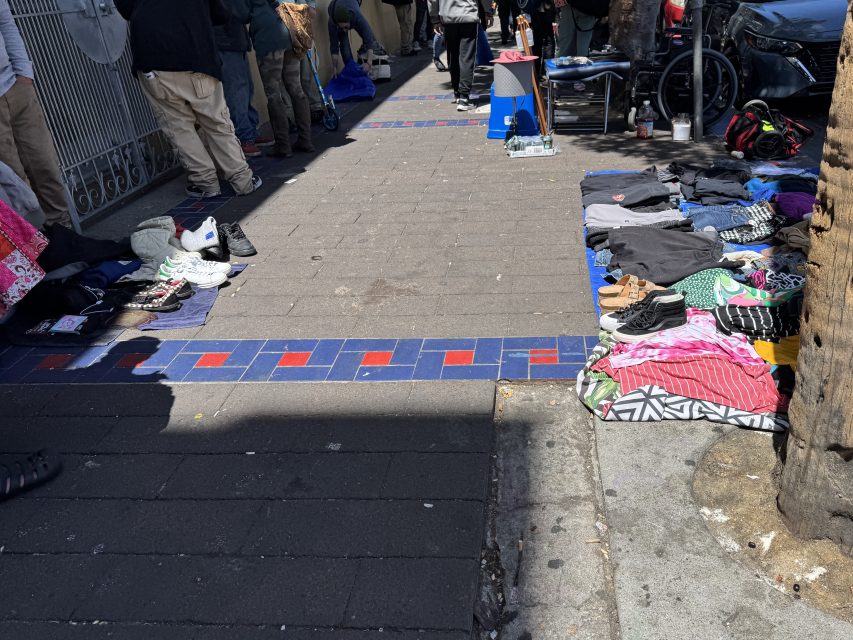 Clothes, shoes, and other items are laid out for sale on the sidewalk, with people standing and walking in the background.