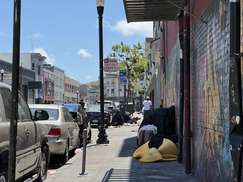A city sidewalk with parked cars, scattered trash, several people sitting or lying down, and graffiti on a nearby wall.