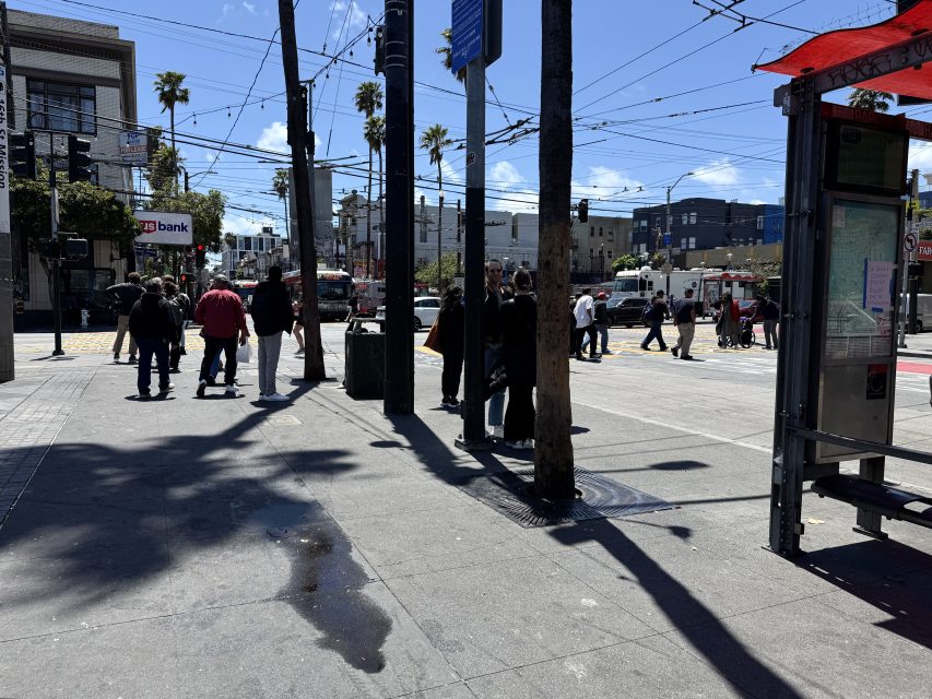 People stand and walk along a city sidewalk near a bus stop on a sunny day, with buildings, palm trees, and a bank in the background.