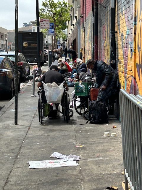 People with belongings, some in wheelchairs, sit and gather on a city sidewalk lined with trash and graffiti-covered walls.