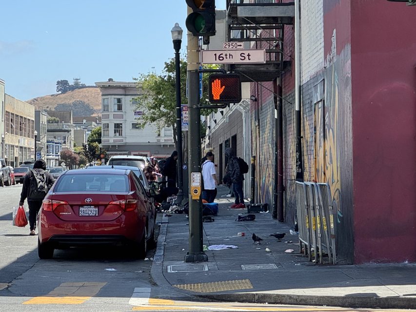 A city street corner at 16th Street with people gathered on the sidewalk, parked cars, a crosswalk signal, and scattered debris on the ground.