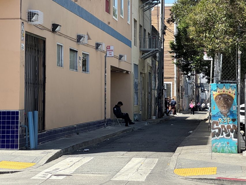 A person sits on a chair in the shade near a building in an alleyway, with a mural and people further down the alley.