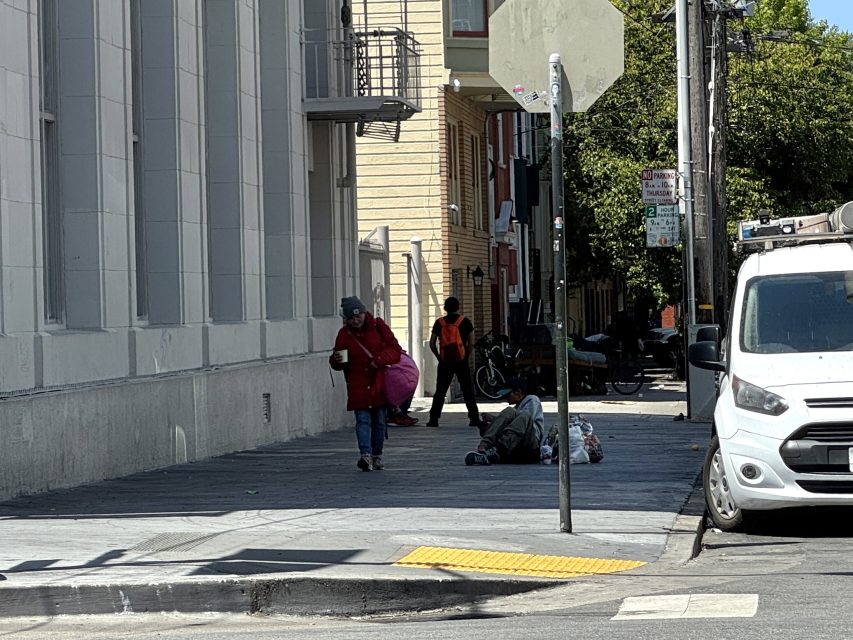 A person walks down a city sidewalk carrying bags, while another person lies on the ground nearby; a white van is parked at the curb.