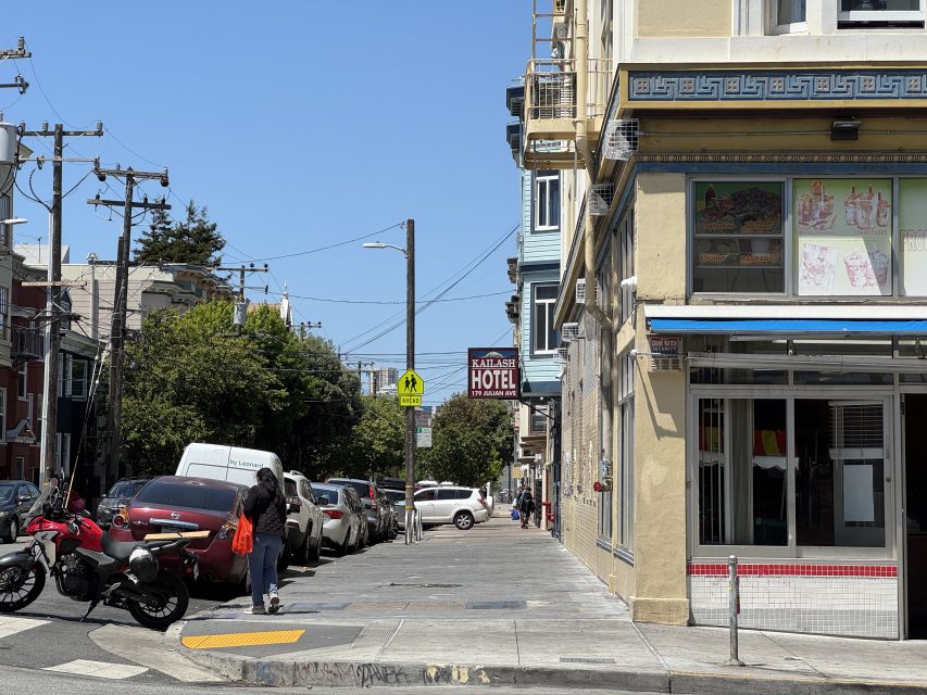 A city sidewalk with parked cars, a motorbike, a few pedestrians, and a building with a "Katalase Hotel" sign in the background on a sunny day.