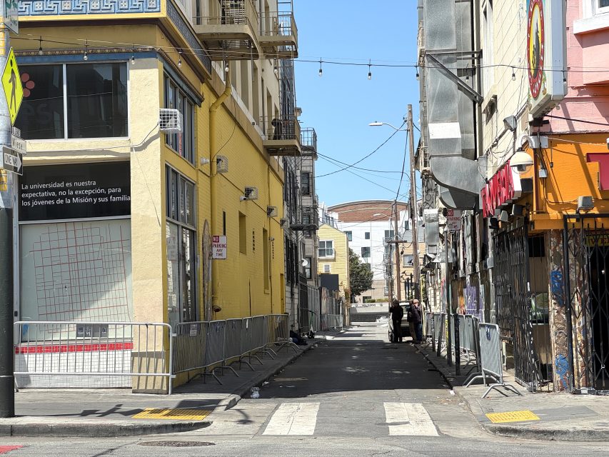 A narrow city alley with yellow and pink buildings, temporary fencing, street art, and some debris on the ground under clear daylight.