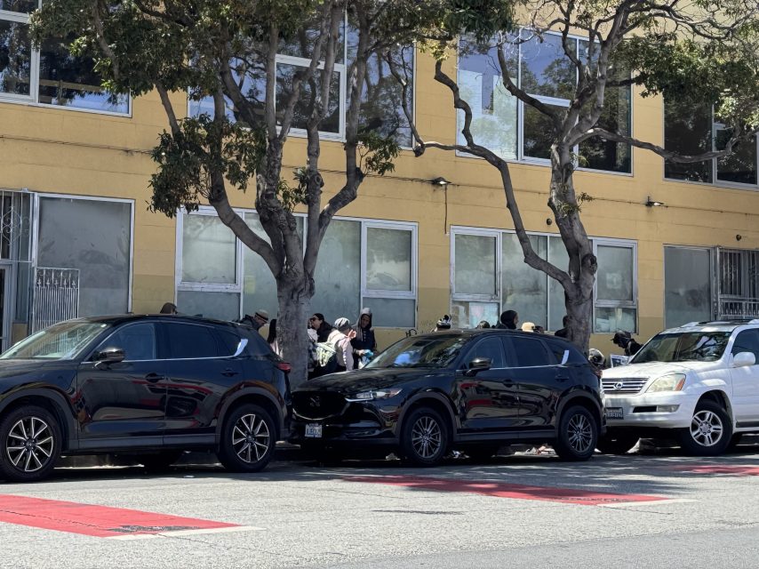 A group of people stands on the sidewalk near a yellow building, partially obscured by parked cars and trees.