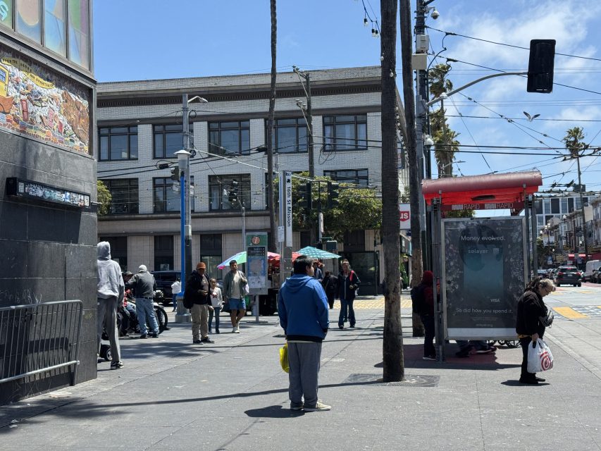 People stand and walk near a city intersection on a sunny day; a bus stop, street art, and several buildings are visible in the background.