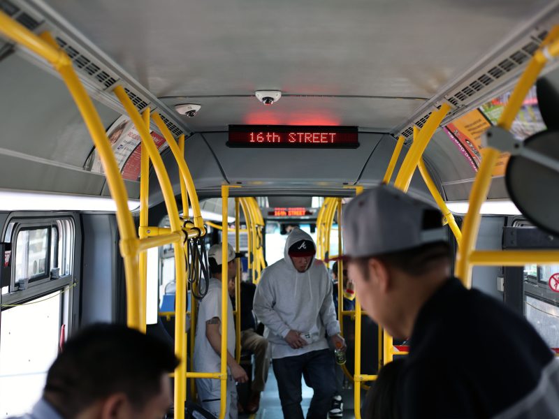 Interior of a city bus with several passengers standing and sitting; an electronic sign displays "16TH STREET" above the aisle.