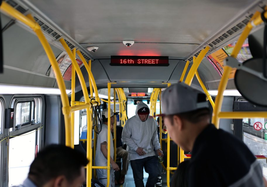 Interior of a city bus with several passengers standing and sitting; an electronic sign displays "16TH STREET" above the aisle.