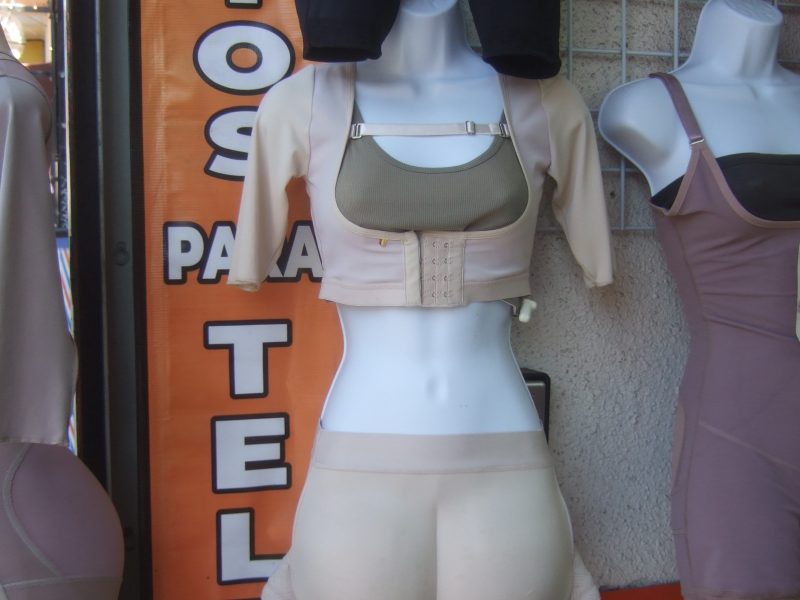 A mannequin displays beige shapewear and a cropped top outside a store, with other shapewear garments hanging nearby and an orange sign in the background.
