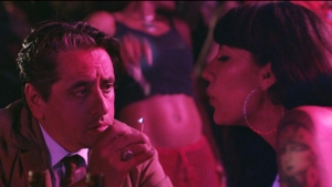 A man in a suit looks intently at a woman with tattoos, who is blowing smoke, in a dimly lit, nightclub-like setting.