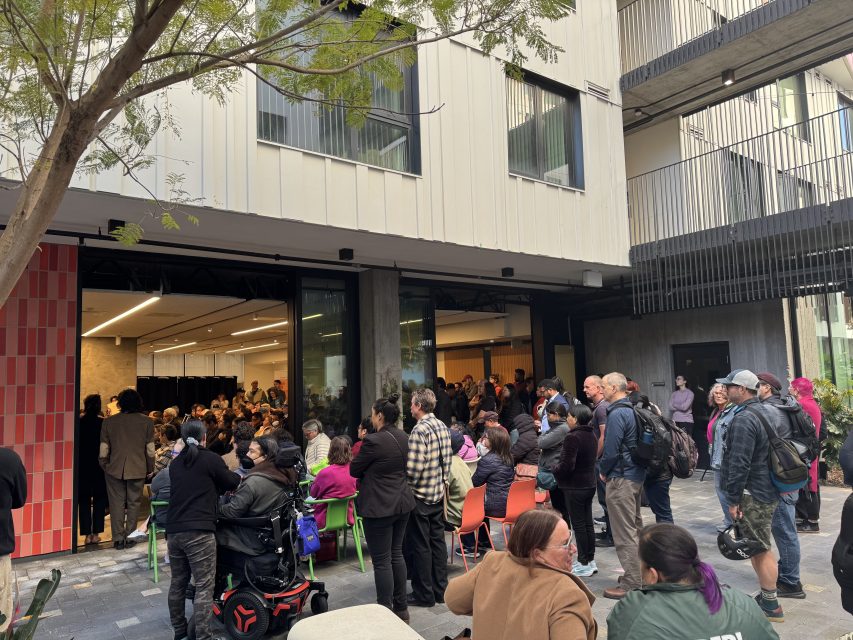A large group of people, some seated and some standing, gather outside and inside a building during what appears to be a public event or meeting.