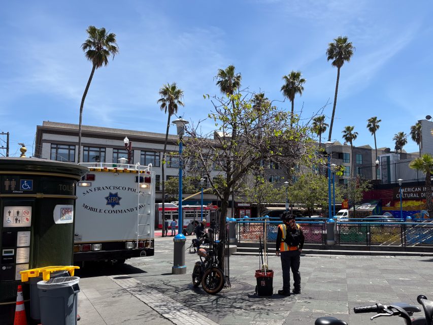 A San Francisco Police mobile command van is parked in a city square near palm trees, bikes, and people on a sunny day.