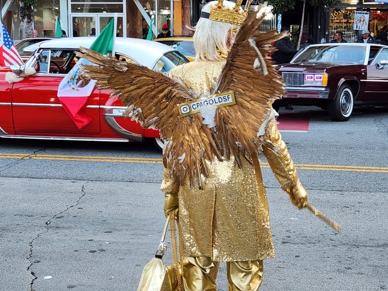 Person in gold sequin outfit and crown with gold angel wings stands on street near red car decorated with flags; a sign reading “@CPAGOLDSF” is attached to the wings.